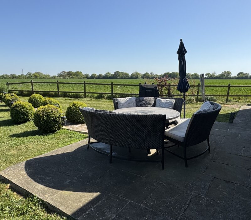 French doors open out onto the patio area. A nine seater, circular table and bench chairs take centre stage. A large blue tilting parasol is folded up, to the right. Beyond this are views across arable farmland, with a row of trees on the horizon.