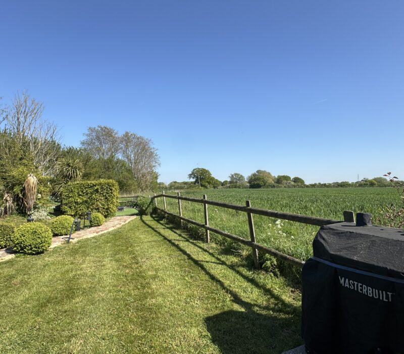 This view from the Shire Stables patio takes in the views across the arable farmland, extending to a row of trees in the distance. There is a large black BBQ and the fence behind it leads to the private hot tub area at the back of the garden. To the left, the new undercover bar area is nestled under trees,