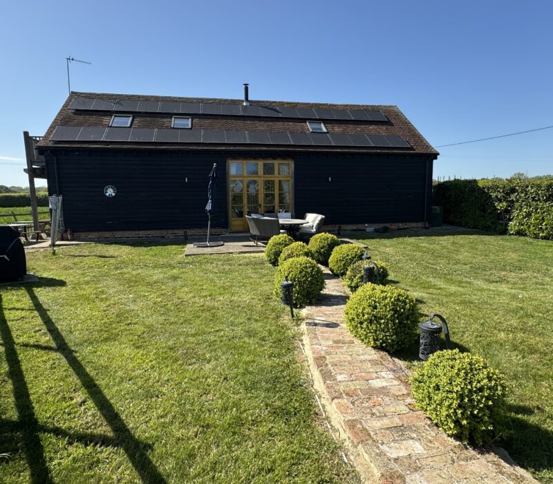 From the back of the garden where the hot tub can be found, a curved brick path, lined either side with large privet balls and solar lamps leads you back to the patio area of the Shire Stables. The large, black, ship lapp timber framed barn contrasts against the green grass in the garden and the clear blue sky.