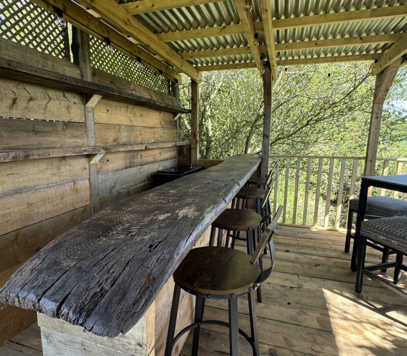 A view inside the bar area. To the right is a high table with four stools. Behind this, the view extends across fields. On the left the barn is constructed from an antique plank of timber and has 4 wooden and metal barn stools underneath. Behind the bar, on the wall, are 2 shelves which are handy for storage. Also behind the bar is a large drinks trolley and cooler that can be filled ice.